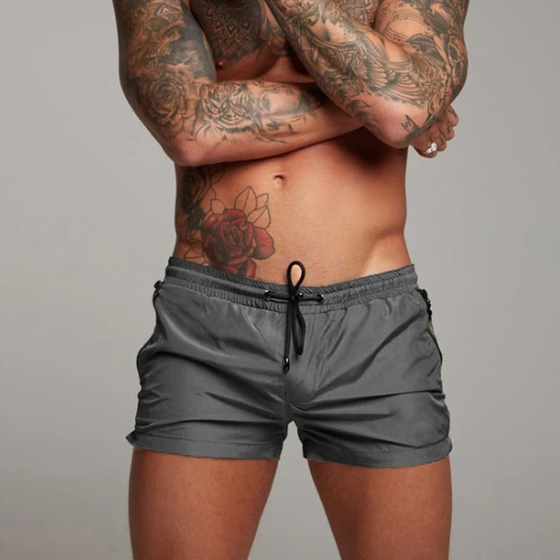 Mann mit tätowiertem Oberkörper trägt graue Badehose. Fitness, Mode, Herrenbekleidung, Sommer, Strandmode.