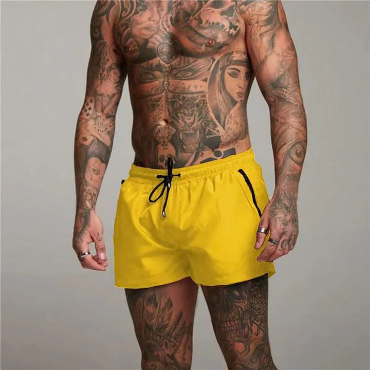 Mann mit tätowiertem Oberkörper trägt gelbe Badehose. Fitness, Mode, Sommer, Strandbekleidung, Herrenmode, Tätowierungen, Sportbekleidung.