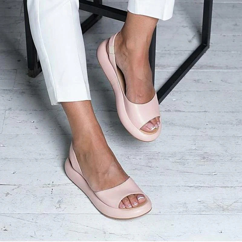 Damenfüße in eleganten, rosa Peeptoe-Ballerinas auf weißem Holzboden, kombiniert mit weißen Hosen. Modische Sommerschuhe für Frauen.