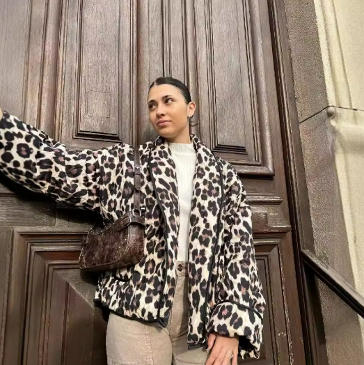 Eine Frau in einer Leopardenmuster-Jacke steht vor einer Holztür. Sie trägt eine braune Tasche und helle Hose. Modetrend, Streetstyle, Wintermode.