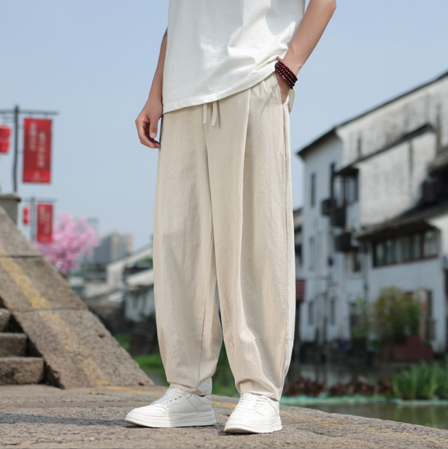 Mann in beiger Leinenhose und weißem T-Shirt steht auf einer Brücke in traditioneller asiatischer Stadt. Lässige Mode, Sommeroutfit, urbane Kulisse.