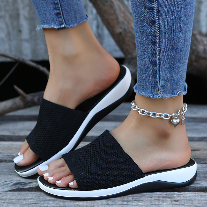 Damenfüße in schwarzen Mesh-Sandalen mit weißer Sohle, kombiniert mit blauen Jeans und silbernem Knöchelarmband, auf Holzboden.