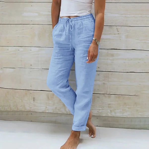 Frau in hellblauer Leinenhose und weißem Top vor Holzwand. Sommermode, lässiger Stil, bequeme Damenbekleidung, Freizeitlook.