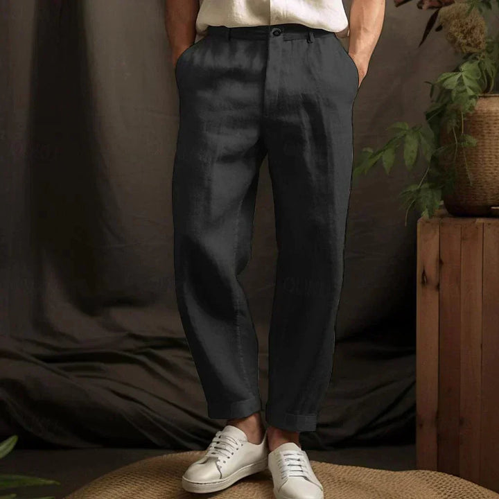 Mann trägt schwarze Leinenhose und weiße Sneaker, steht vor einem braunen Hintergrund mit Pflanzen. Elegante Herrenmode, lässiger Stil.