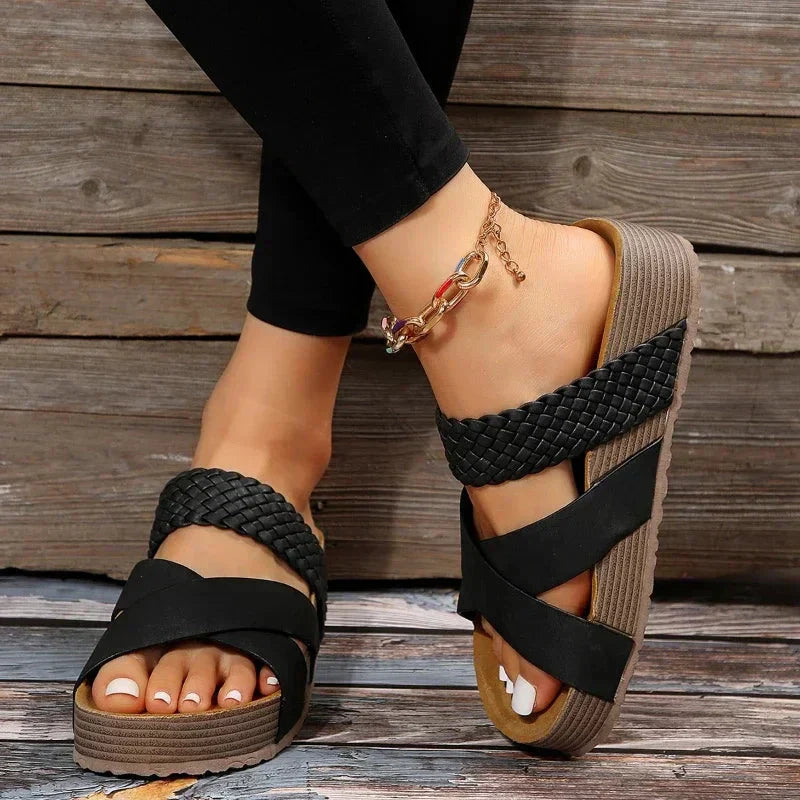 Schwarze geflochtene Sandalen mit Plateau, getragen von einer Person mit Fußkettchen, vor Holzhintergrund. Modische Damen-Sommerschuhe.