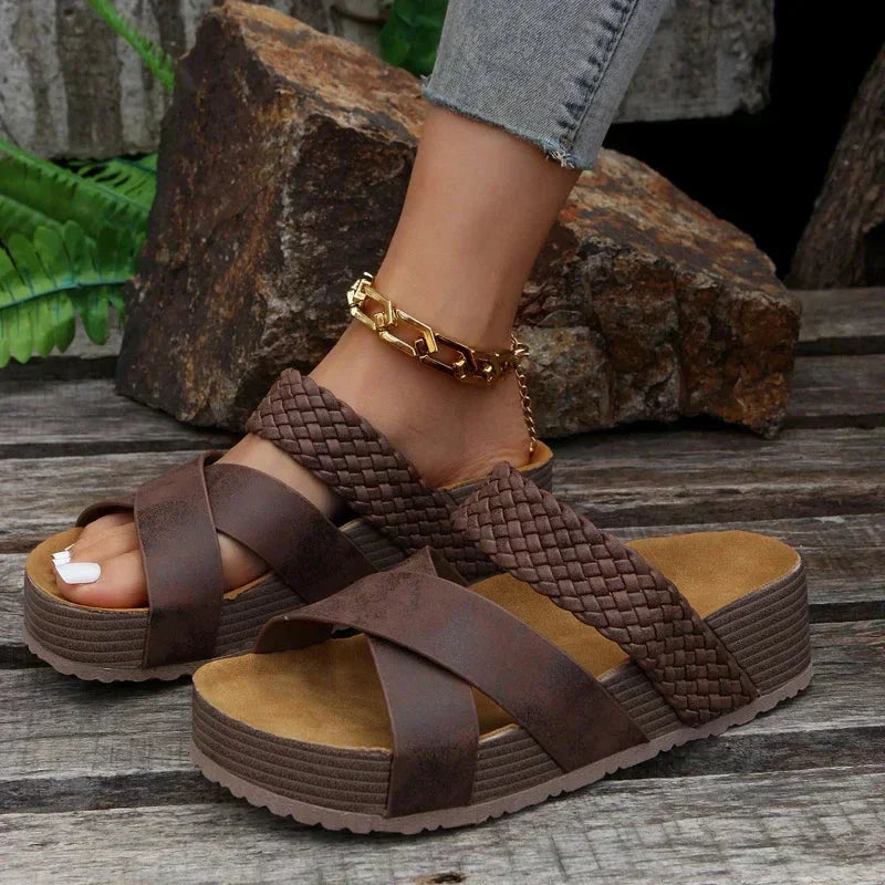 Damenfuß in braunen, geflochtenen Plateau-Sandalen auf Holzuntergrund, mit goldener Fußkette. Modische Sommerschuhe, bequem und stilvoll.