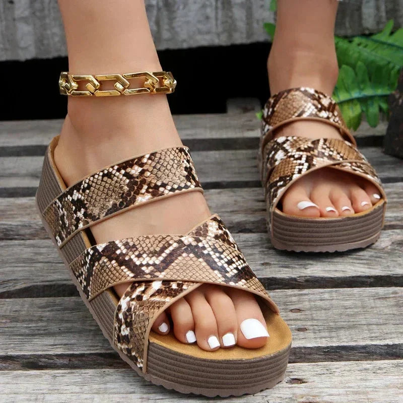 Damen Sandalen mit Schlangenmuster, Plateau-Sohle und goldener Knöchelkette auf Holzuntergrund. Modische Sommerschuhe, trendiges Design.