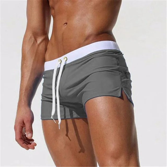 Mann in grauer Badehose mit weißem Bund und Kordelzug, muskulöser Oberkörper, Strandhintergrund, Sommermode, Herrenbademode.