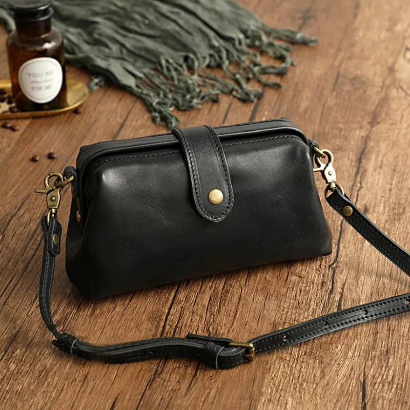 Schwarze Ledertasche mit Schnalle und abnehmbarem Schulterriemen auf Holztisch, neben grünem Schal und Glasflasche. Vintage-Stil, elegant.